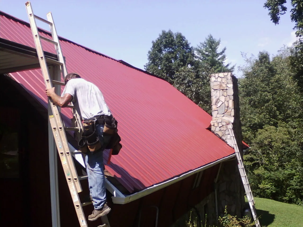Roofer on a ladder beside a red metal roof and stone chimney — Soffit & Fascia Repair by Roofing Brothers in Quebeck